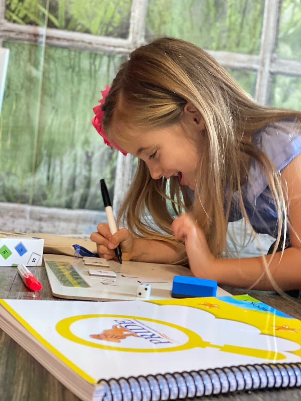 A smiling girl leans over a whiteboard to write with the PRIDE Reading Program Yellow Workbook in the foreground