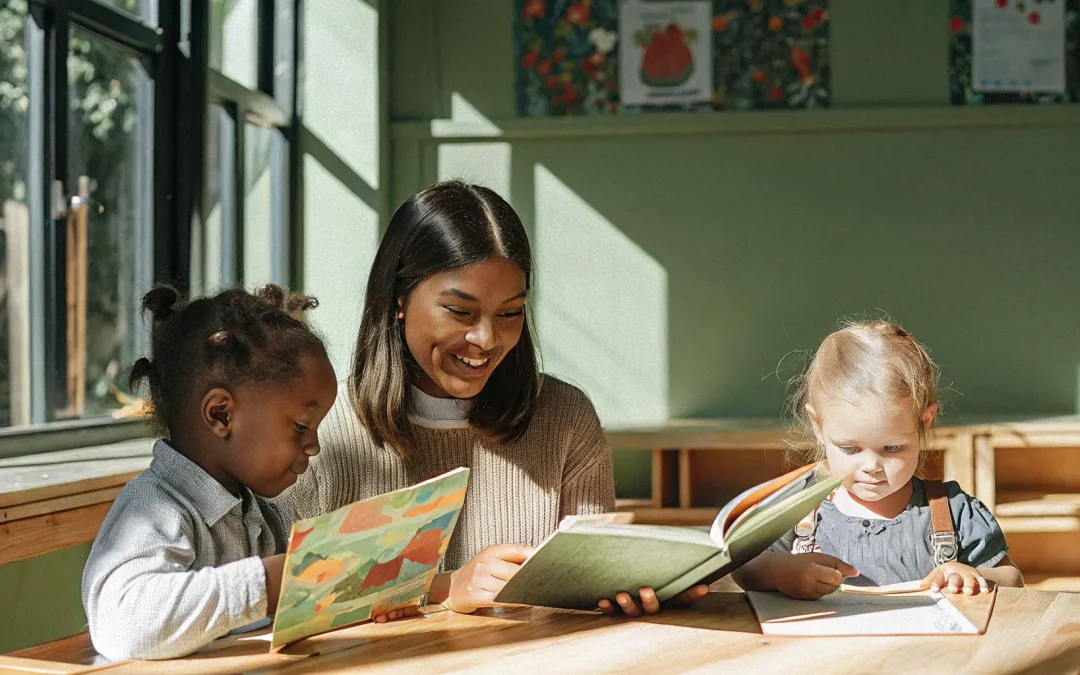 A beautiful, bright, professional real classroom photograph of a smiling female teacher sitting at a wooden table with a diverse group of elementary children, guiding them as they read colorful decodable books in a sunlit classroom